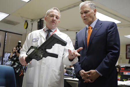 Photo of Gov. Jay Inslee being shown a semi-automatic rifle fitted with a bump stock.