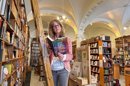 a woman in glasses stands on a ladder reading a book in a book store