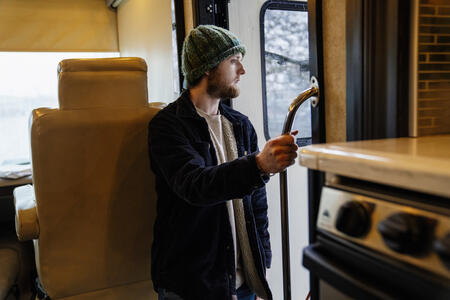 A man, wearing a beanie and fleece-lined coat, looks to the outside of an RV.
