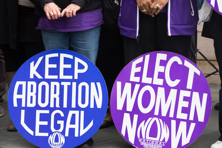 A blue sign says "Keep Abortion Legal" and a purple sign says "Elect Women Now." 