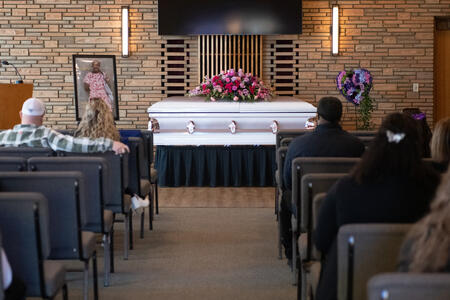 People in chairs look at a coffin at the front of a room decorated with flowers