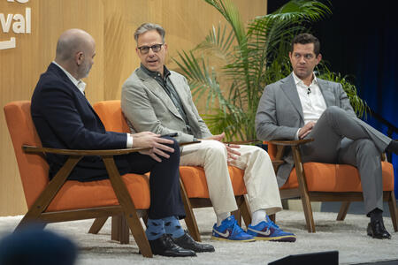 Three people in suits sit on orange chairs on stage with plants in the background. The two people on the right are looking at the person on the left, who is looking back at them.
